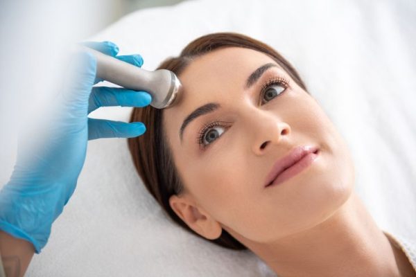 Skin care and treatment. Close up top angle portrait of relaxed smiling brunette lady having rejuvenation roll massage of forehead in spa center making by therapist